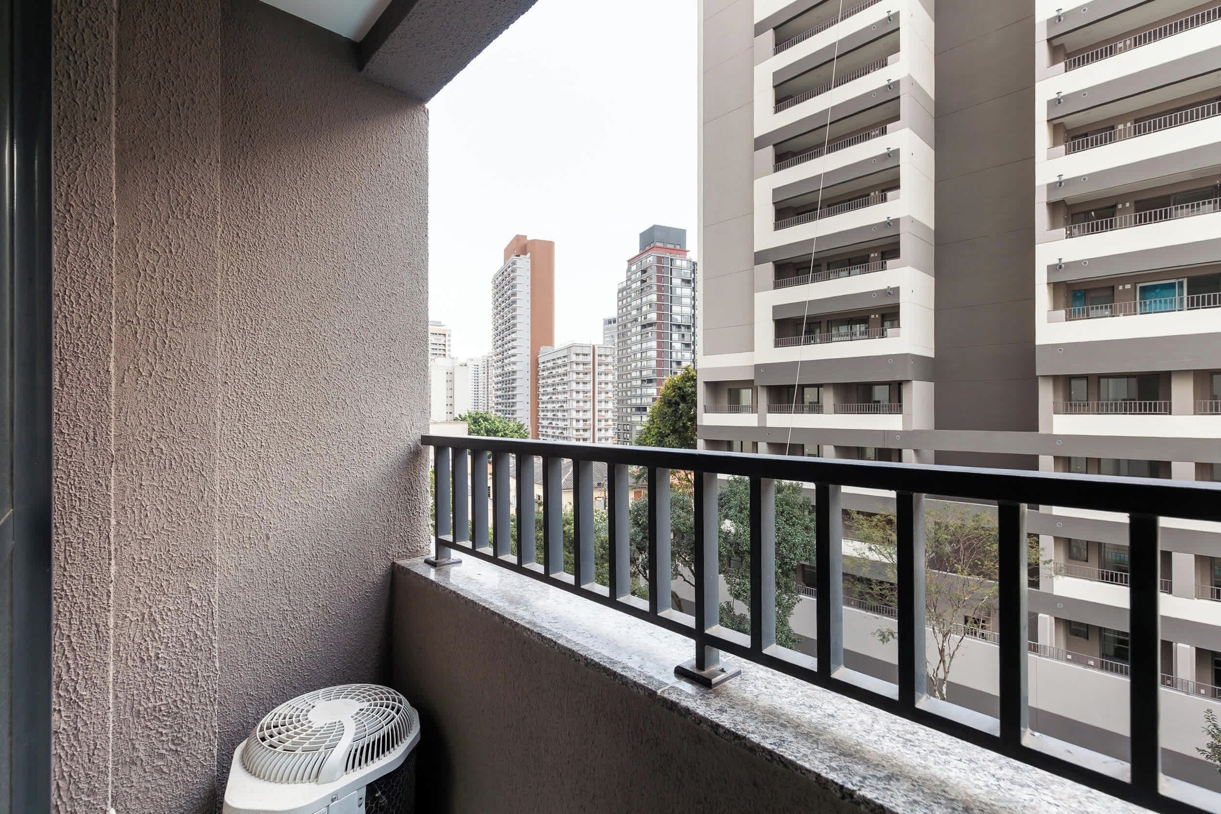 A balcony with a metal railing, a trash bin, and a view of high-rise buildings in the background.