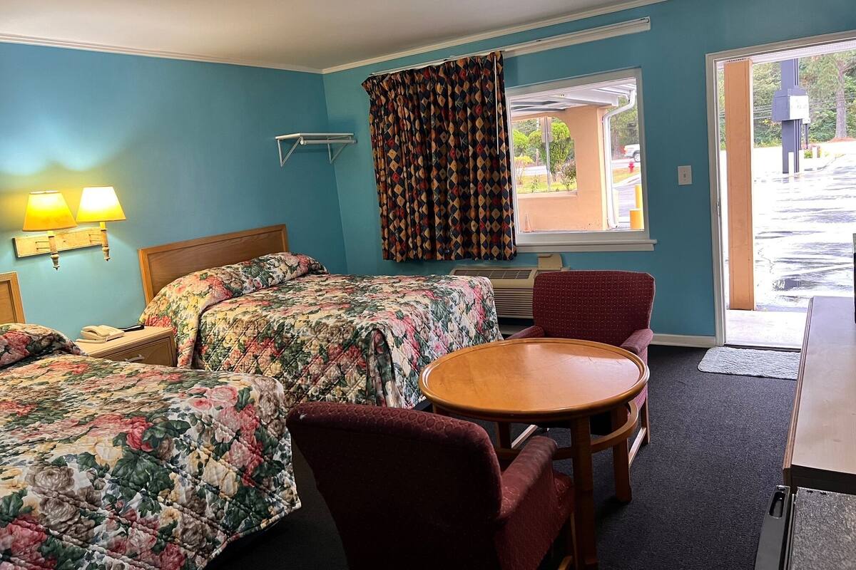 A hotel room with two beds, a small table, a chair, a window with curtains, and a view of the outside.