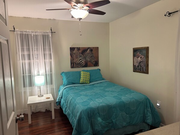 A bedroom with a bed, a ceiling fan, a window with sheer curtains, and two wall-mounted paintings.