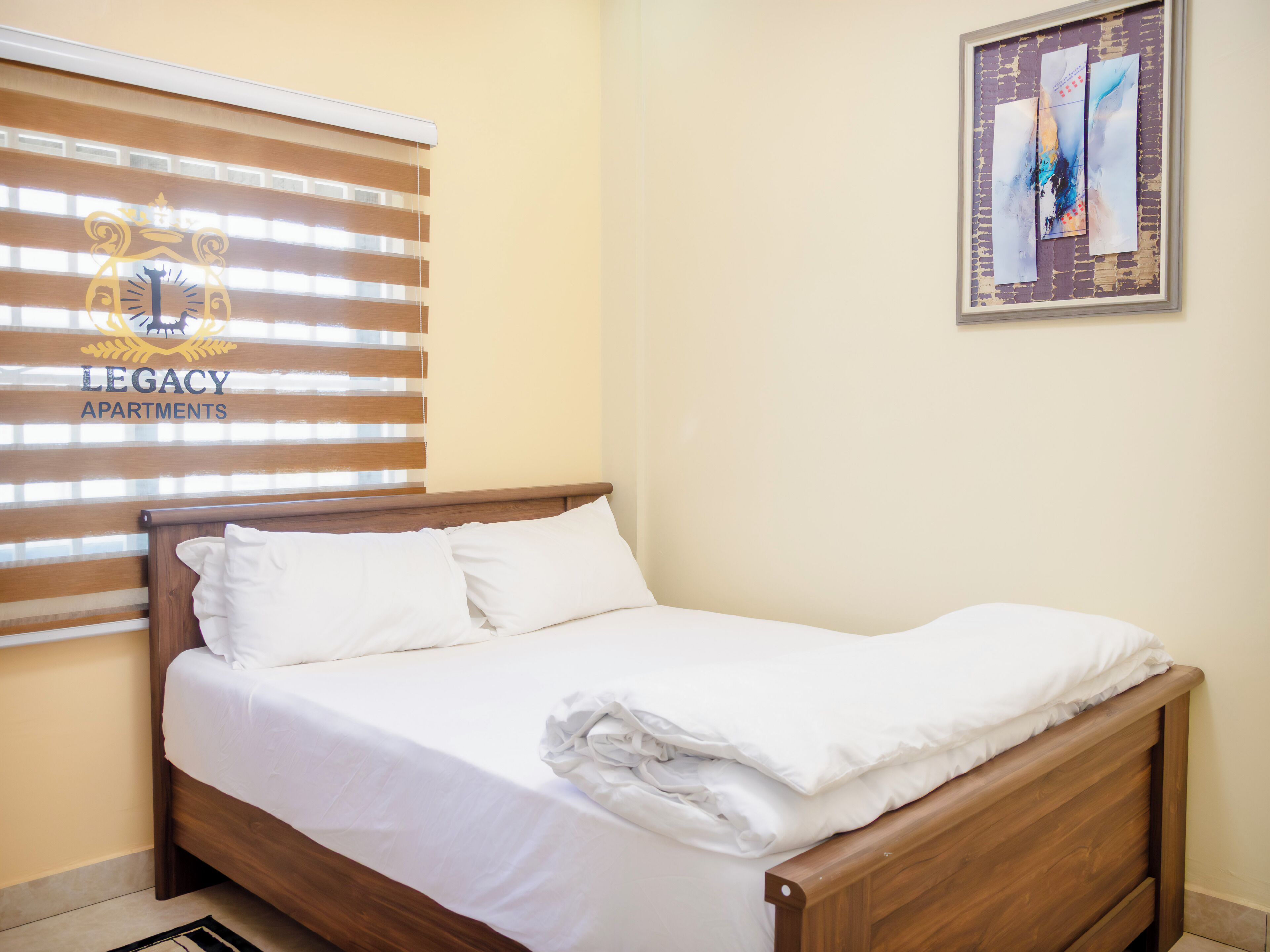 A neatly made bed in a room with a wooden headboard, a window with blinds, and a framed abstract painting on the wall.