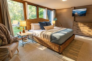 A bedroom with a large bed, a TV, a window with a view of trees, and a wicker chair.