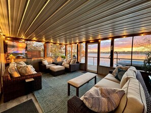 A spacious indoor area with a large window overlooking a body of water, furnished with wicker sofas and a flat-screen TV.