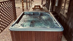 A hot tub under a wooden pergola with string lights, surrounded by a wooden deck and a picnic table.