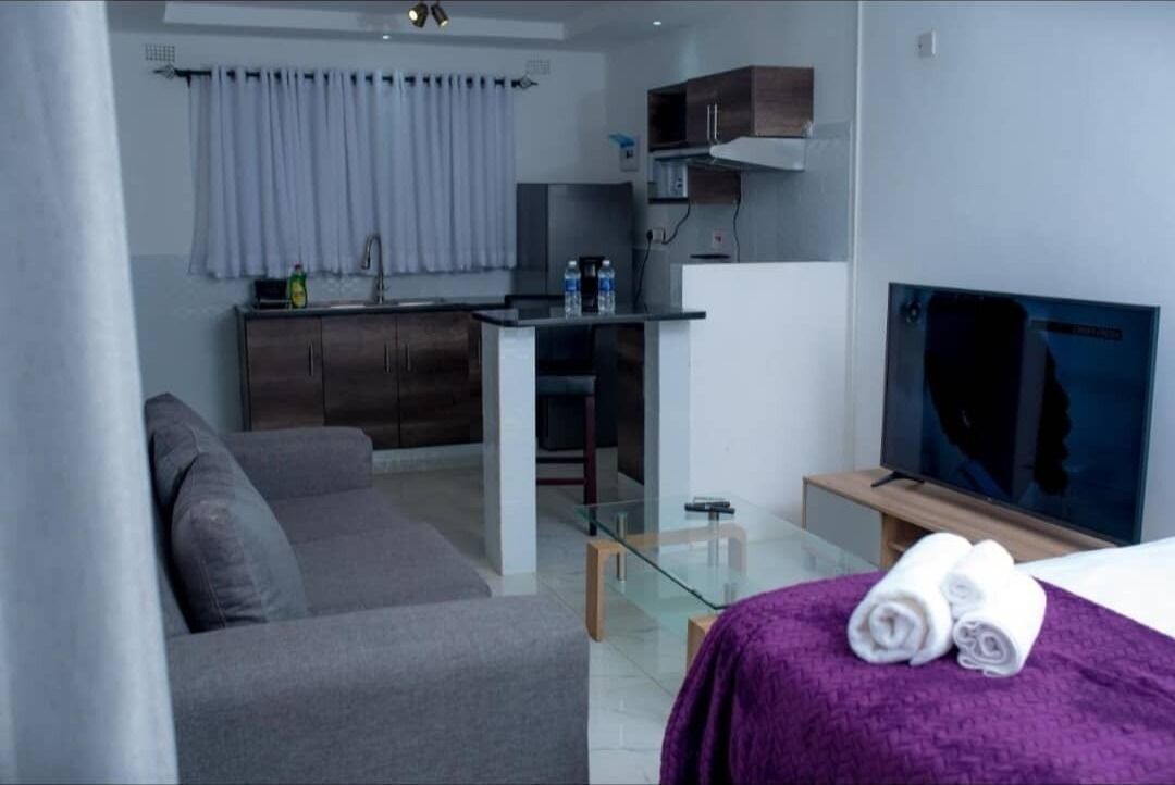 A modern hotel room with a grey sofa, a small kitchen area with a microwave, a glass table, and a TV on a wooden stand.