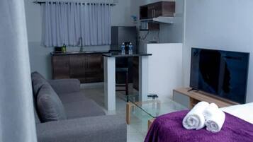 A modern hotel room with a grey sofa, a small kitchen area with a microwave, a glass table, and a TV on a wooden stand.