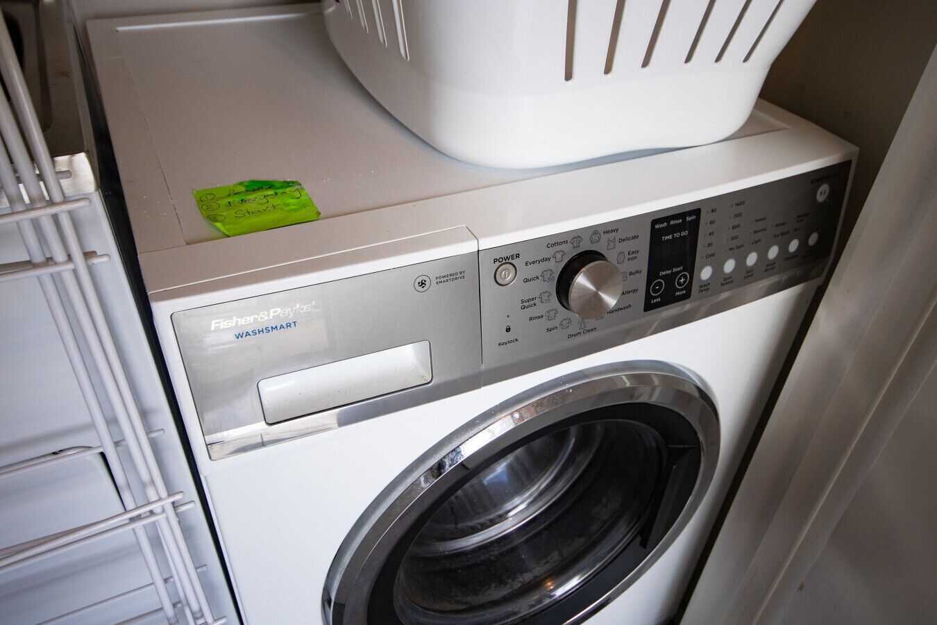 A front-loading washing machine with a control panel, a green note, and a white basket on top.