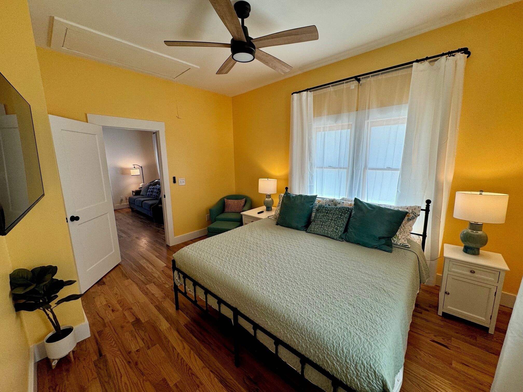 A bedroom with a bed, a ceiling fan, a nightstand, a lamp, a window with curtains, a door, and a plant.