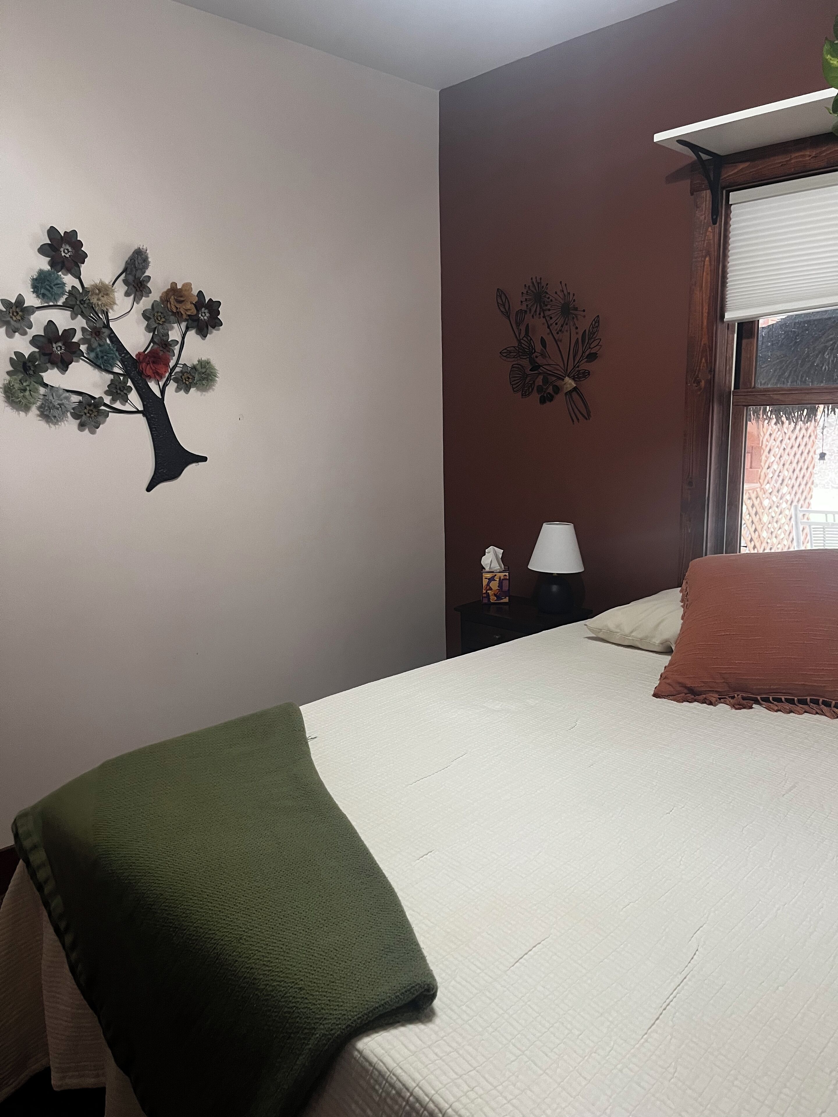 Una camera da letto con un letto, un comodino con una lampada, una finestra con persiane e decorazioni murali tra cui un albero e un motivo floreale.