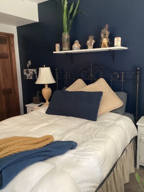 A bedroom with a bed, a nightstand, a lamp, a shelf with decorative items, and a door.