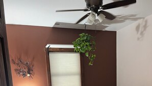 Un dormitori amb un llit, una tauleta de nit amb una làmpada, un ventilador de sostre i una planta penjant.