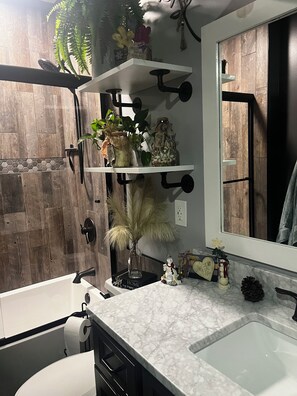 A modern bathroom with a white sink, a mirror, and a shower. There are shelves with plants and a small figurine.