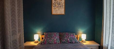 Een slaapkamer met een donkerblauwe muur, een bloemenprint bedspreidek, twee bedlampen en een ingelijst kunstwerk boven het bed.