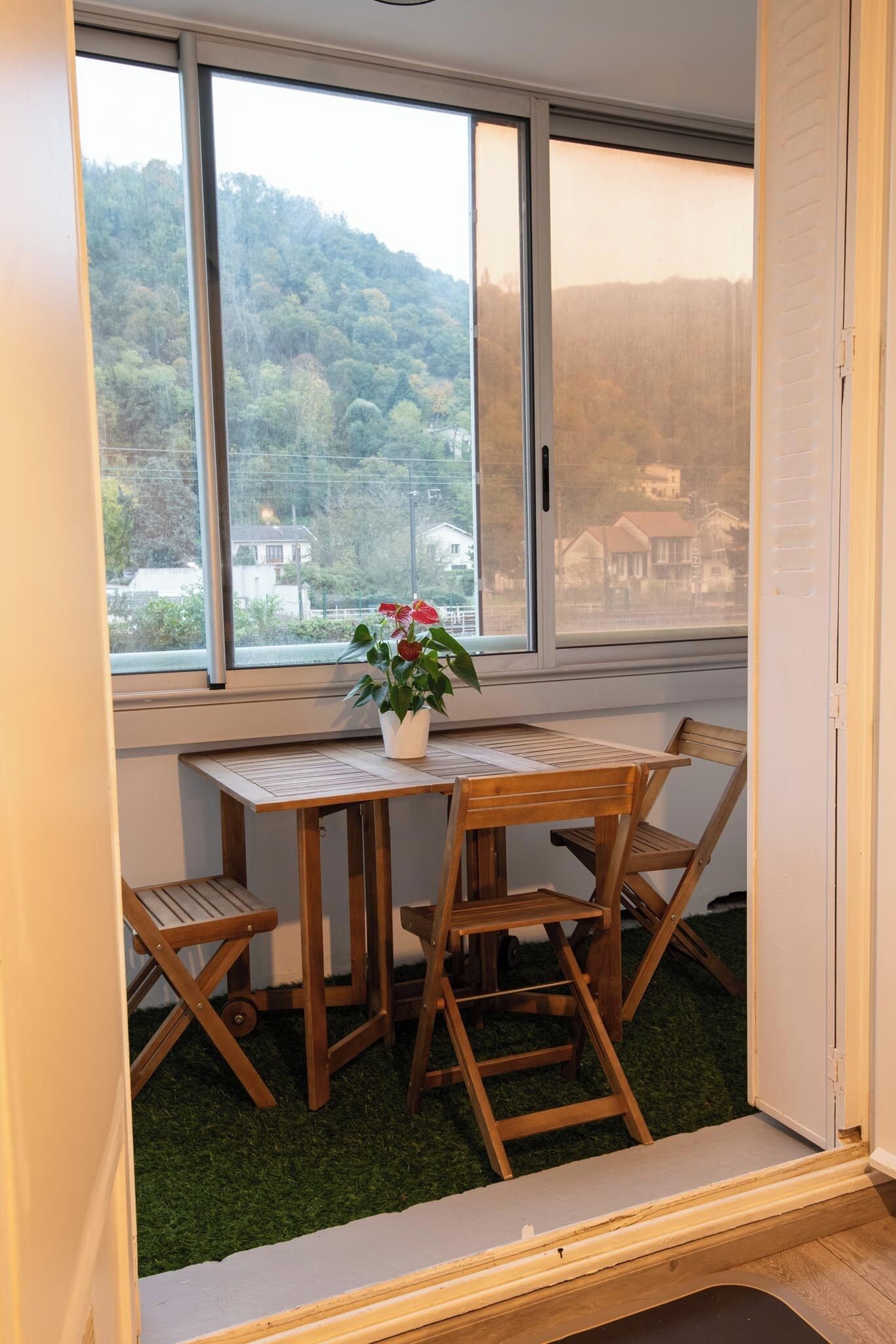 A corner room with a wooden table and chairs, a potted plant, and a view of a forested hill through a large window.