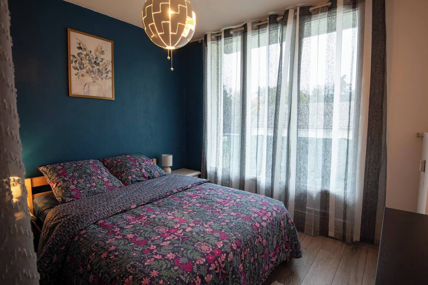Une chambre à coucher avec un lit, une table de chevet, une lampe, un tableau et une fenêtre avec des rideaux.