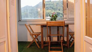 Een hoekkamer met een houten tafel en stoelen, een raam met uitzicht op een beboste heuvel, en een kleine bloempot op de vensterbank.