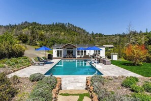 Une maison avec une piscine, un patio doté de chaises longues et une vue sur une colline boisée.