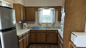 A compact kitchen with wooden cabinets, a stainless steel refrigerator, a built-in oven, and a microwave on a wooden cart.