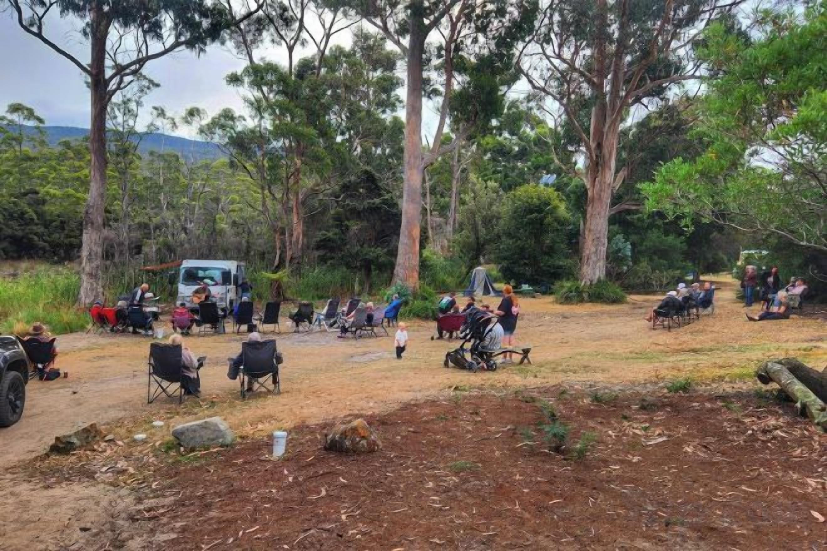 An outdoor gathering with people seated on chairs and camping gear, surrounded by trees and a dirt ground.