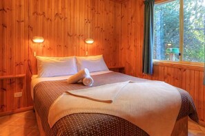 A wooden-paneled room with a bed, two wall-mounted lights, and a window with a view of trees. - Bruny Island Holiday Park Adventure Bay (Adventure Bay)