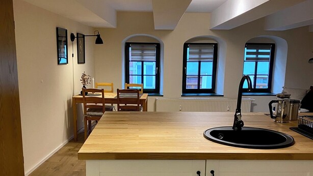 A modern kitchen with a central island, white cabinetry, and a dining area with a wooden table and chairs. - 1.5-room apartment with terrace (Pirna)