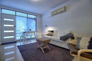 A modern living room with a white sofa, a small wooden table, and a large window with blinds.