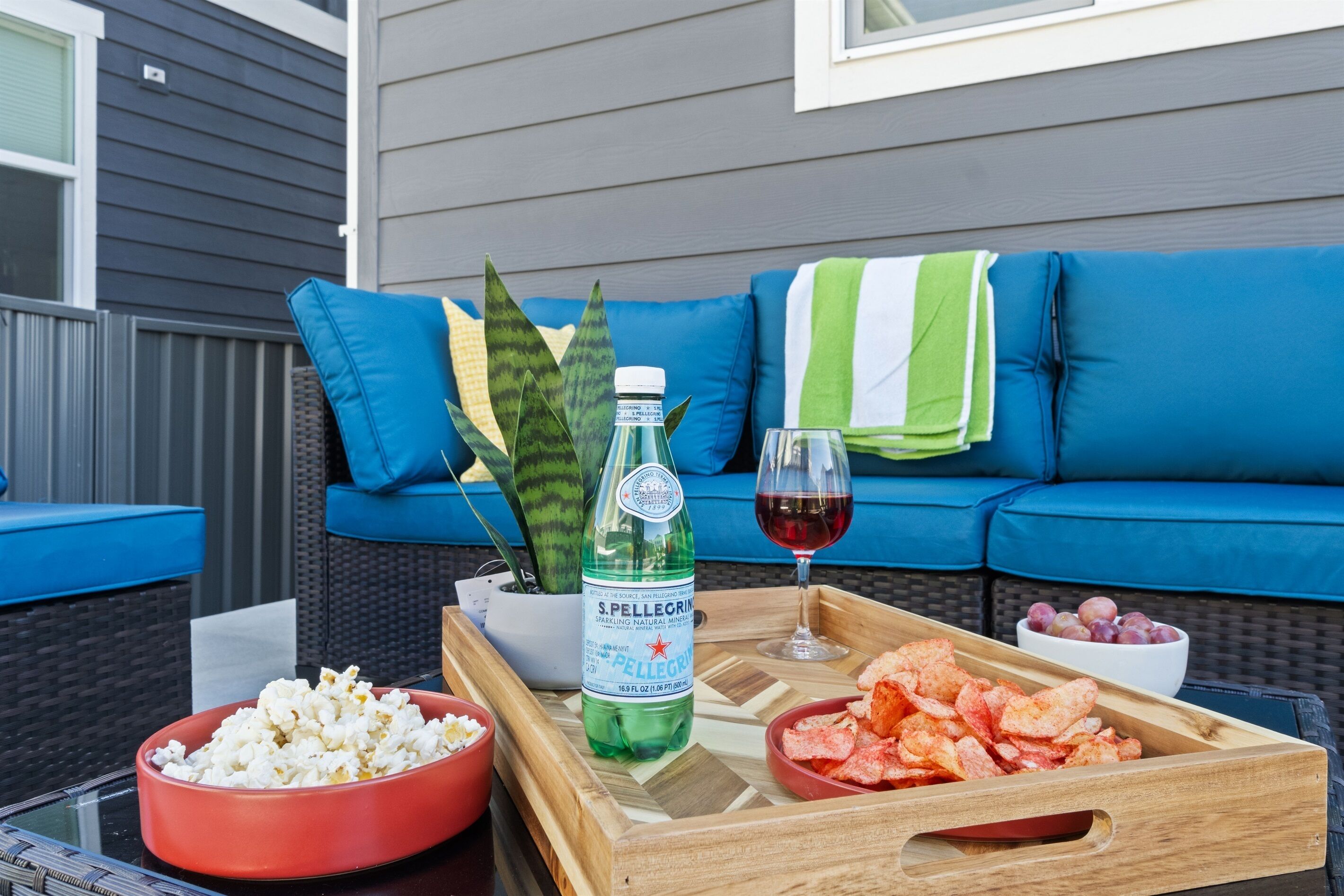 Ein Holztablett mit Popcorn, Chips, einer Flasche Sprudelwasser und einem Glas Rotwein auf einem Tisch vor einer blauen Couch mit einem grün-weißen Streifenhandtuch.