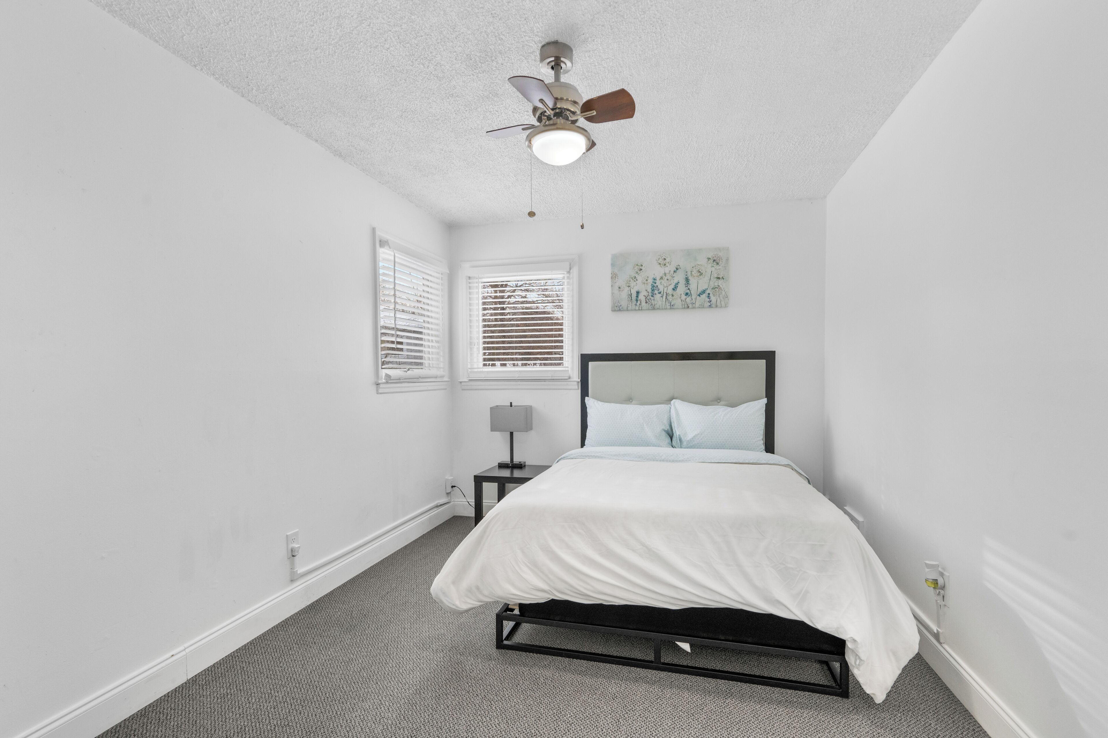 A bedroom with a bed, a nightstand, a ceiling fan, a window with blinds, and a wall-mounted artwork.