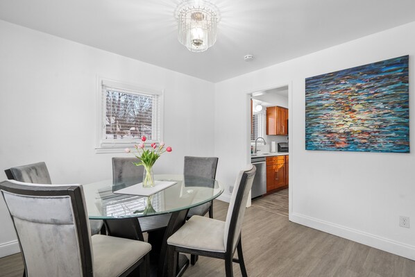 Un espace de salle à manger moderne avec une table en verre, des chaises rembourrées, un vase de fleurs, un lustre, une fenêtre avec des stores et un tableau abstrait coloré accroché au mur.