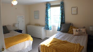 Une chambre avec deux lits, un ventilateur de plafond, une fenêtre avec des rideaux, une commode et une lampe de chevet.
