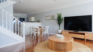 A modern living room with a wooden dining table, white chairs, a large TV, and a staircase. - The Jetty Retreat - 1km from Jetty Beach (Coffs Harbour)