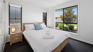 A modern bedroom with a large bed, a nightstand, a window with a view of greenery, and a door. - Moonee Beach Getaway - (Moonee Beach)