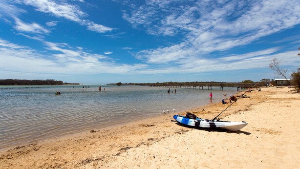 A beach with a wooden pier extending into calm waters, a boat on the shore, and people engaging in water activities. - Myee Court - 500 meters to Urunga Sea Lido (Urunga)