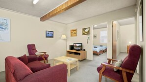 A cozy living room with red upholstered chairs, a wooden coffee table, and a television on a wooden stand. There are framed pictures on the wall and a bed with a blue and white striped blanket in the background. - Myee Court - 500 meters to Urunga Sea Lido (Urunga)