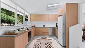 A modern kitchen with wooden cabinets, a stainless steel refrigerator, and a large window offering a view of greenery. - Maison Blanc, Avalon IH (Avalon Beach)