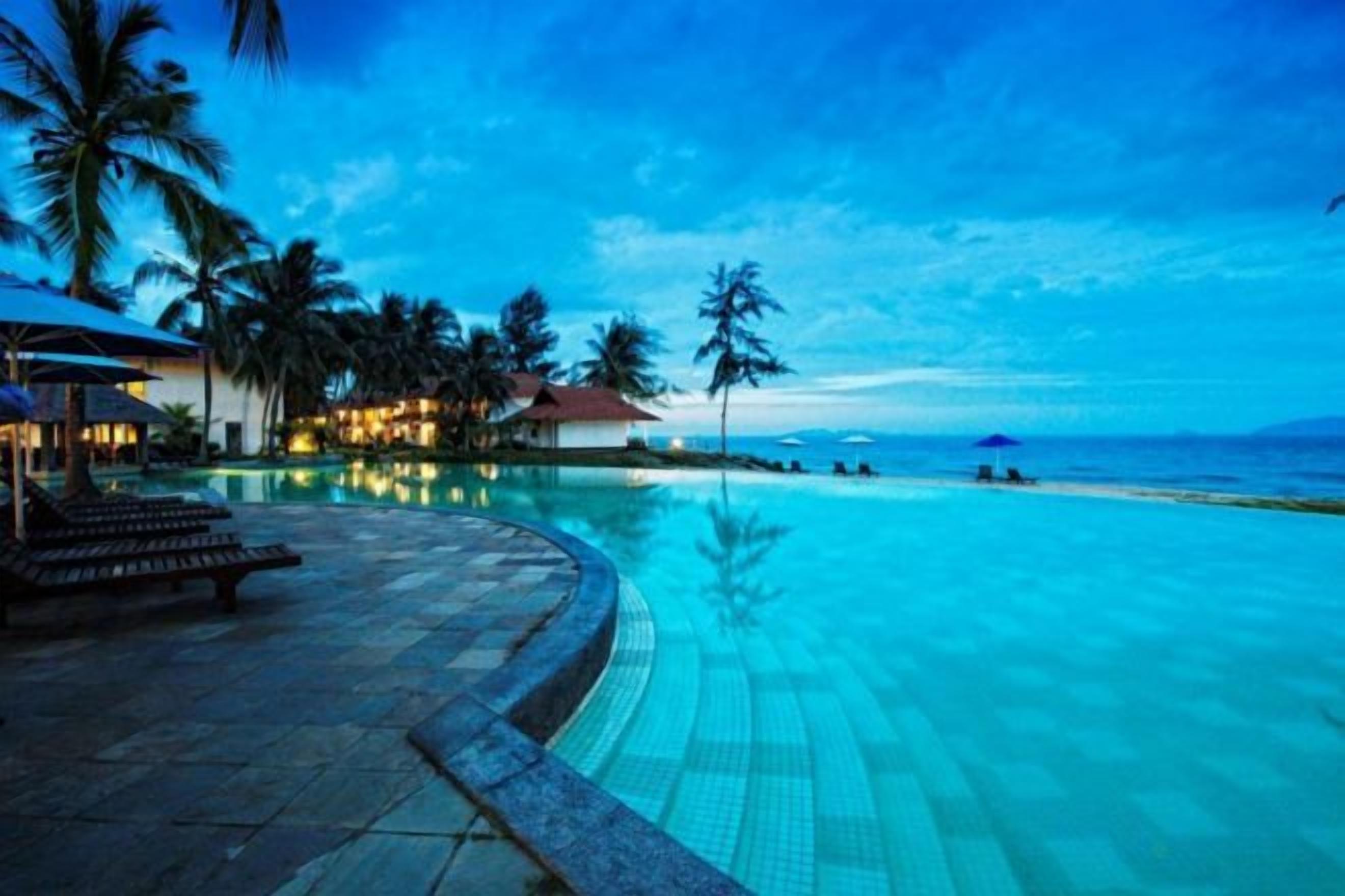 A resort pool with a curved edge, palm trees, and a beach in the background.