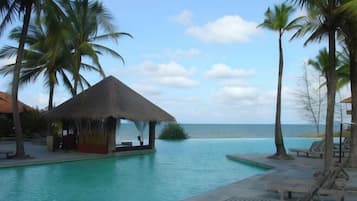 Ein Poolbereich eines Resorts mit einer strohgedeckten Struktur, Palmen und Liegestühlen.