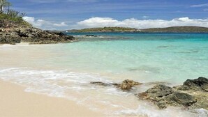 A beach with clear turquoise water, rocky shoreline, and distant hills.