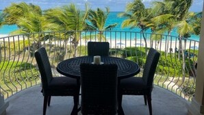 Un balcón con mesa y sillas con vista a una playa con palmeras y cielo despejado.