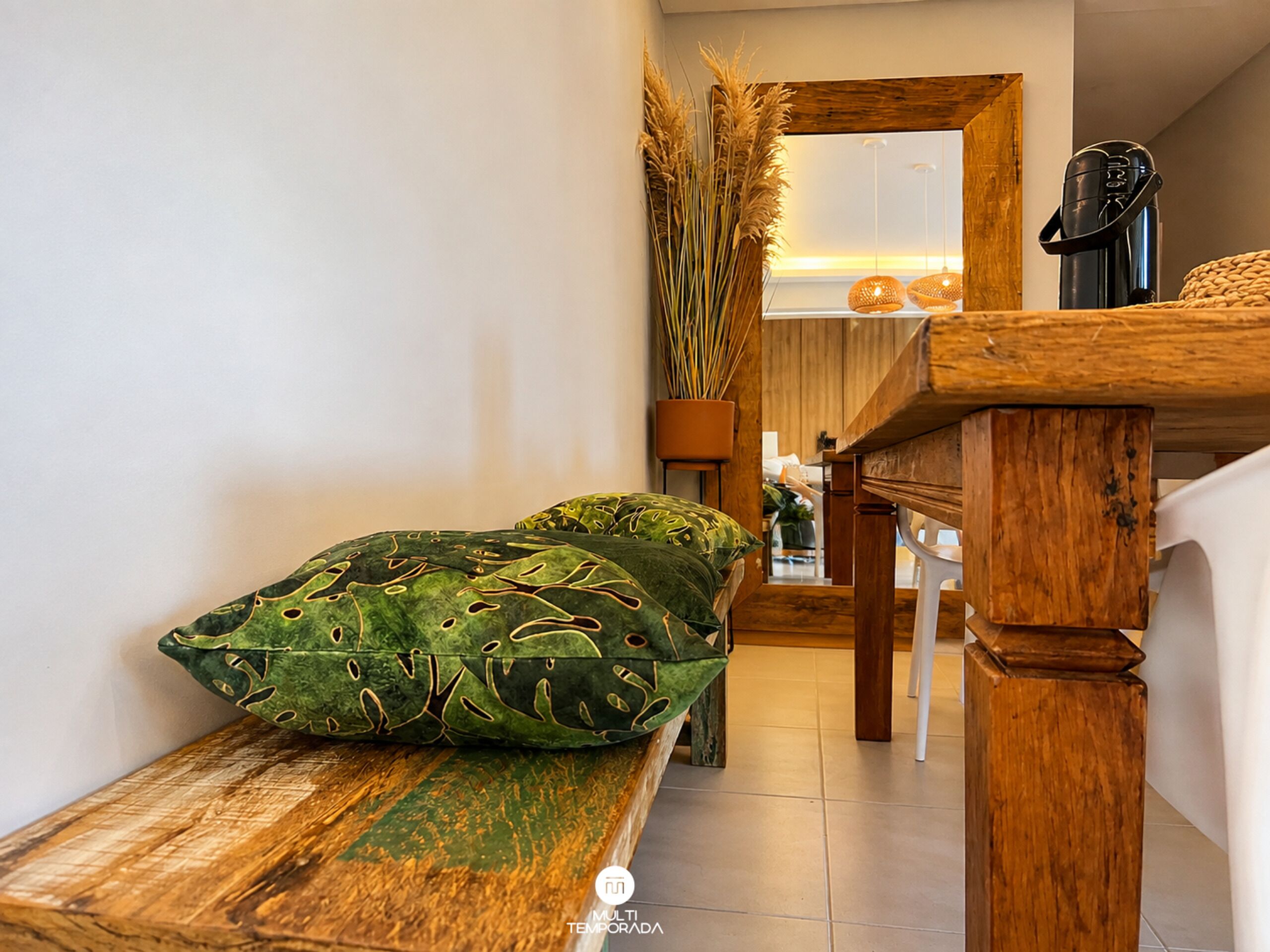 A wooden bench with a cushion, a wooden table, and a mirror reflecting a room with a plant and a coffee machine.