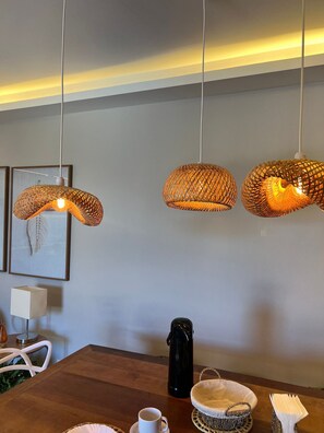 A dining area with a wooden table, three woven pendant lights, a framed artwork, a white lamp, a black coffee pot, and a woven basket.