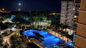 Vista noturna de uma área de piscina de hotel, com piscina circular, espreguiçadeiras e palmeiras.