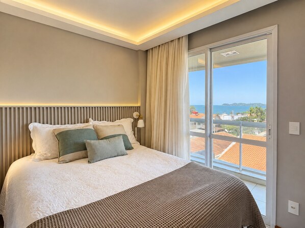 A hotel room with a bed, a nightstand, a lamp, and a view of the sea through a large window. - Casa Refugio Vista Mar - Nautilus Penha (Penha)