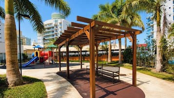 A covered outdoor seating area with a bench, surrounded by palm trees and a playground with a slide.