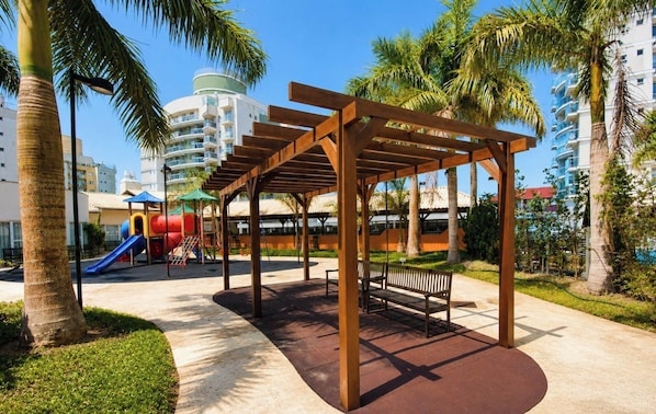 A covered outdoor seating area with a bench, surrounded by palm trees and a playground with a slide. - Casa Refugio Vista Mar - Nautilus Penha (Penha)