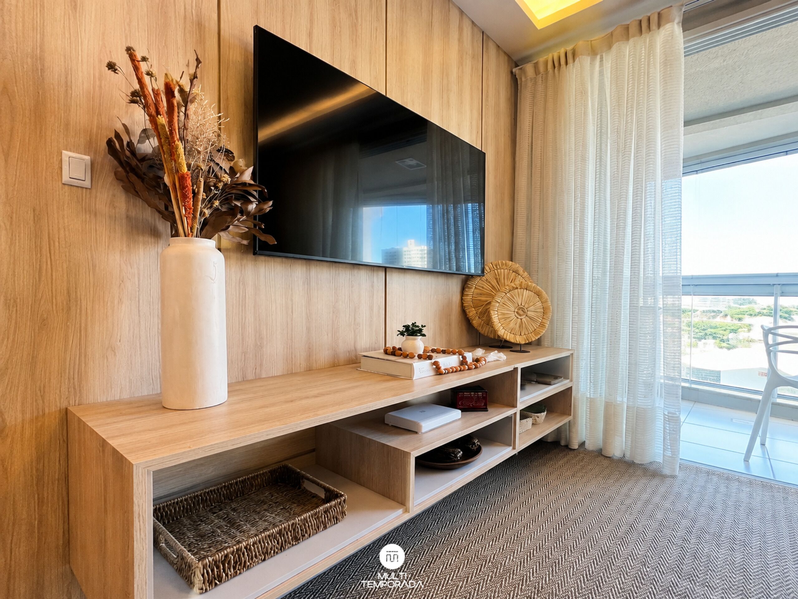 Un salon moderne avec un téléviseur à écran plat fixé à un mur en bois, un vase blanc contenant des plantes séchées, un panier en osier et une petite table agrémentée d’objets décoratifs.