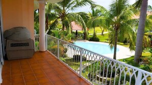 Un balcon donnant sur une piscine entourée de palmiers et d’un jardin.