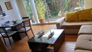 En stue med et glas sofabord, trægulv, en sofa med en gul pude og et spiseområde med et glasbord og stole.