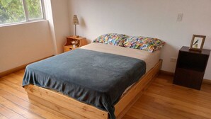 En træseng med et blåt tæppe, en farverig pude, et natbord med en lampe og et lille træskab.