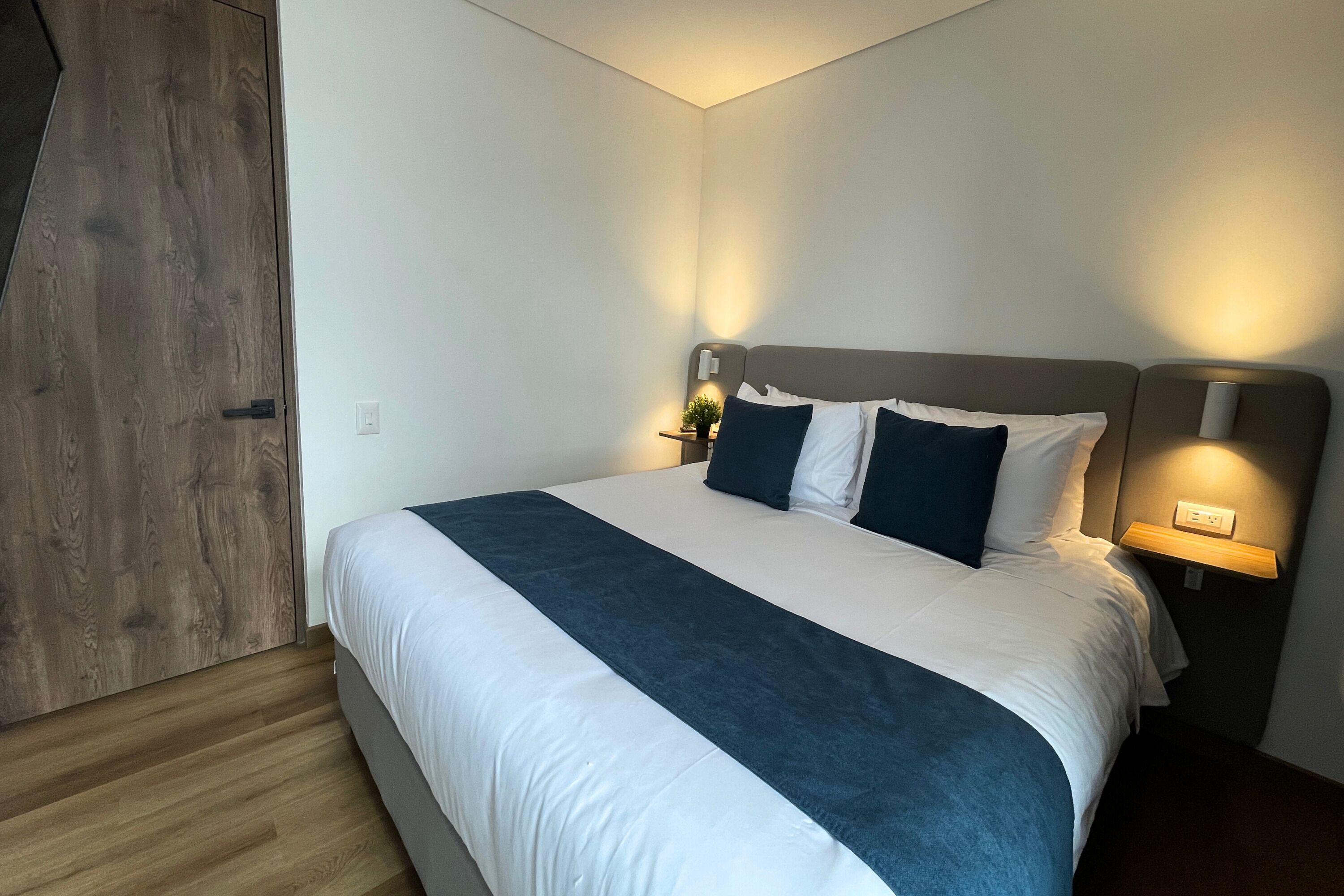A neatly made bed with a white and dark blue bedspread, two bedside lamps, and a wooden wardrobe.
