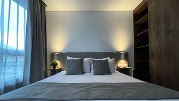 A modern hotel room with a large bed, two bedside tables with lamps, a large window with curtains, and a wooden wardrobe.